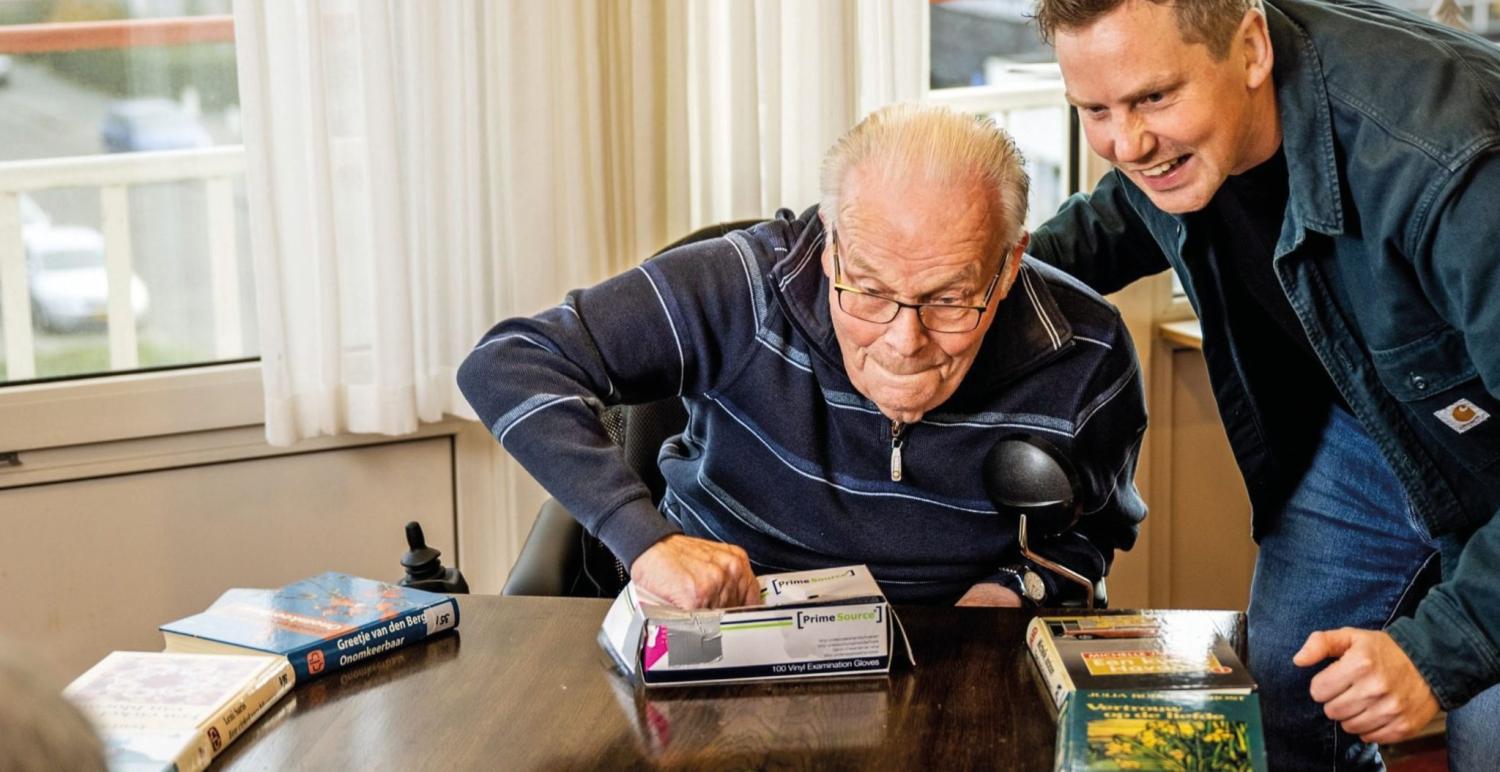 Oudere meneer die een beweegactiviteit doet aan tafel en hierbij geholpen wordt.